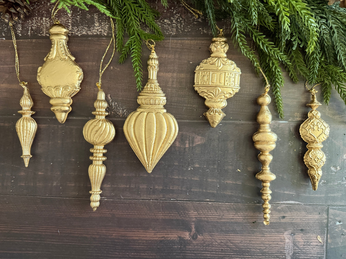 A set of seven vintage-style gold Christmas ornament baubles in various shapes, hanging against a dark rustic wooden background with green pine branches at the top.