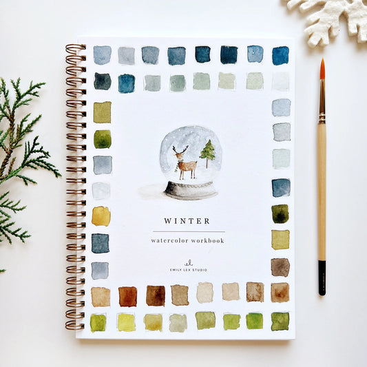 A Winter Watercolor Workbook with colorful illustrations and watercolor brush beside it, on a white background with pine needles in the background.