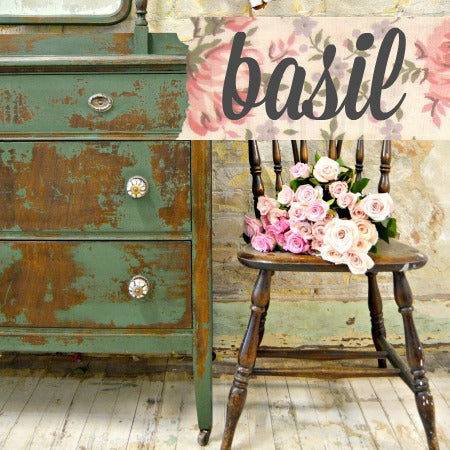 A vintage suitcase painted in dark forest green milk paint alongside a wooden chair with flowers on top, set against a wall with floral wallpaper.