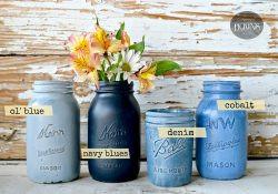 Jars of Sweet Pickins Milk Paint in different shades of blue, placed on a wooden surface with a floral arrangement in the background.