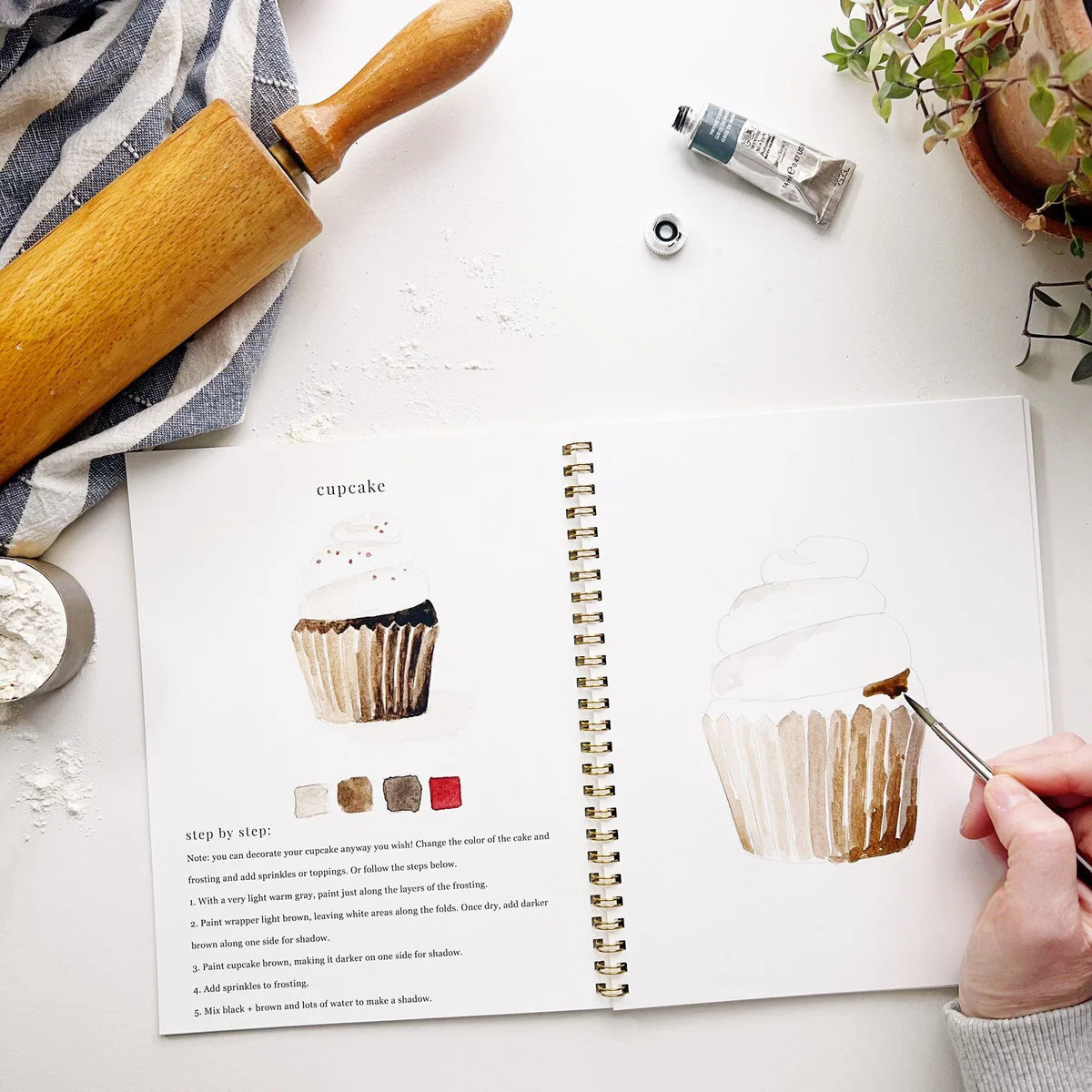 An open watercolor workbook on a white table, with a hand following instructions to paint a cupcake.
