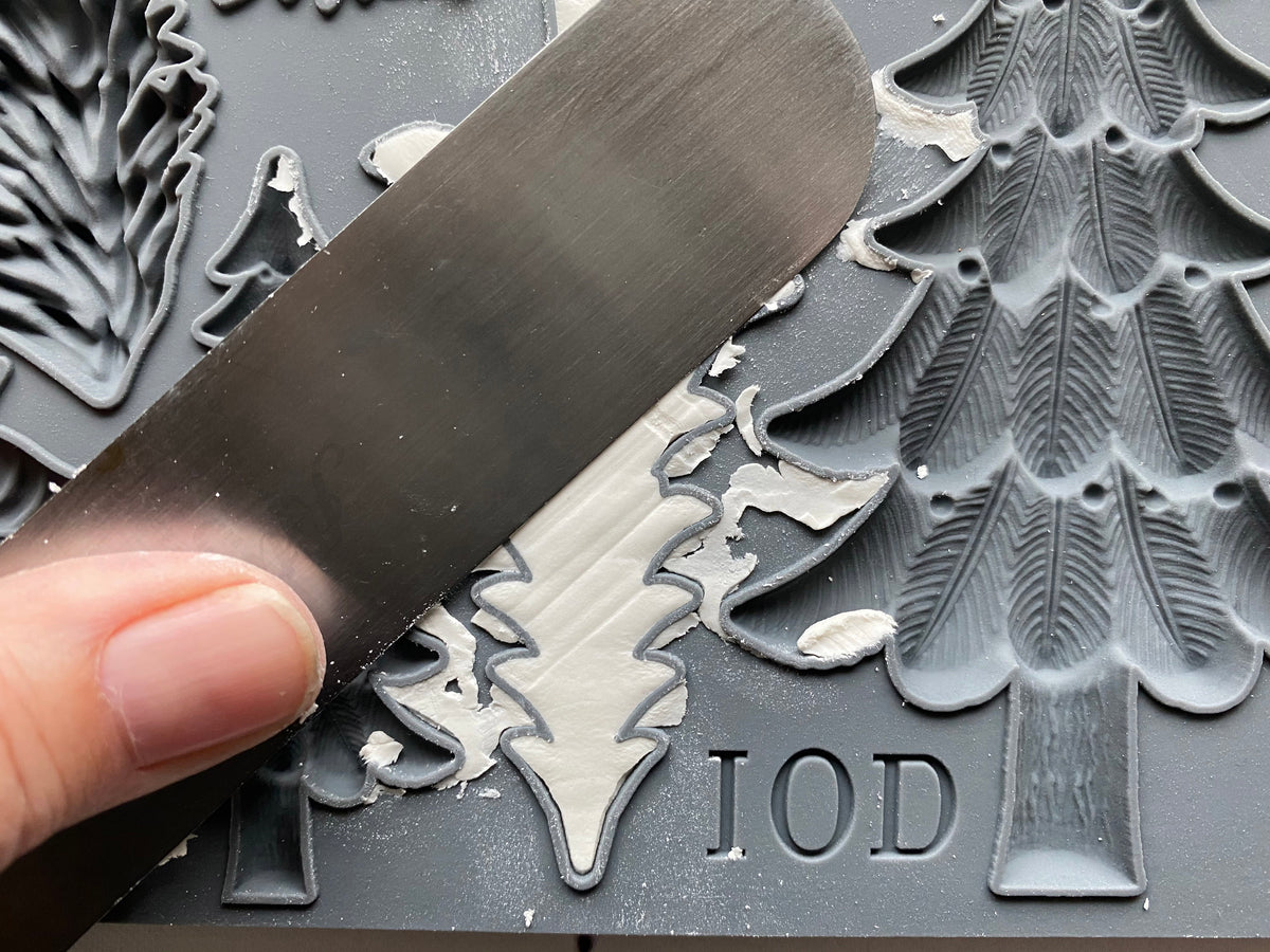 Hand using a metal spatula to scape excwess air dry clay off of the pine design in the IOD mould.