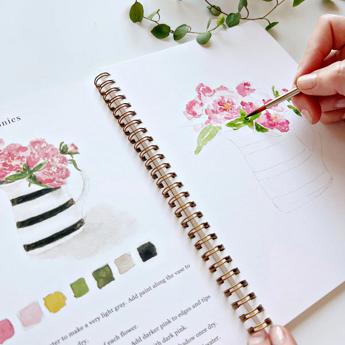 A hand holding a brush painting a bouquet of flowers in a workbook
