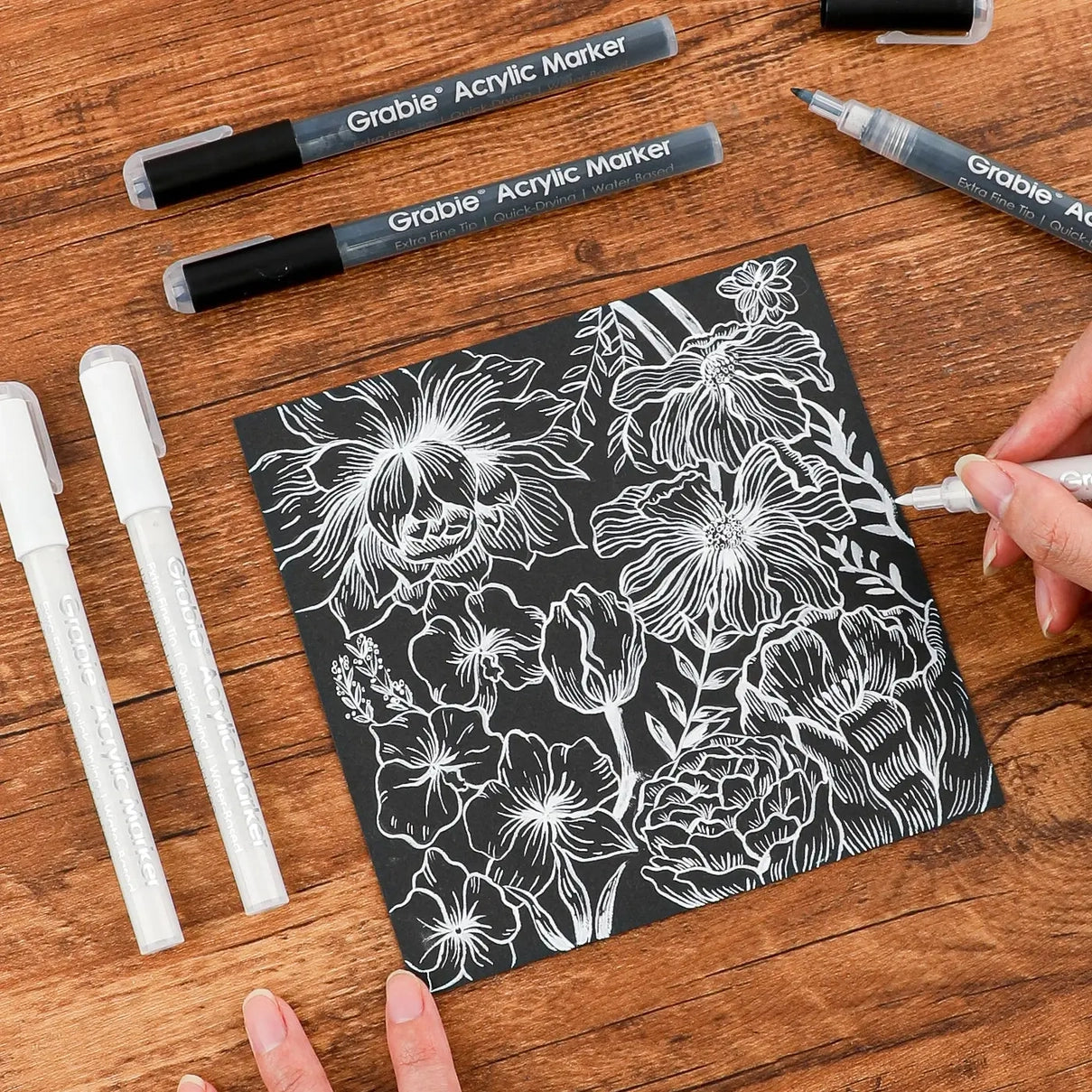Floral design on black paper surrounded by Grabie acrylic markers on a wooden surface
