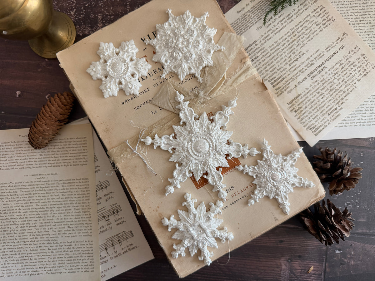 Five white air dried clay snowflakes arranged on an old book, surrounded by vintage pages, pinecones, and a brass object on a dark wooden surface.
