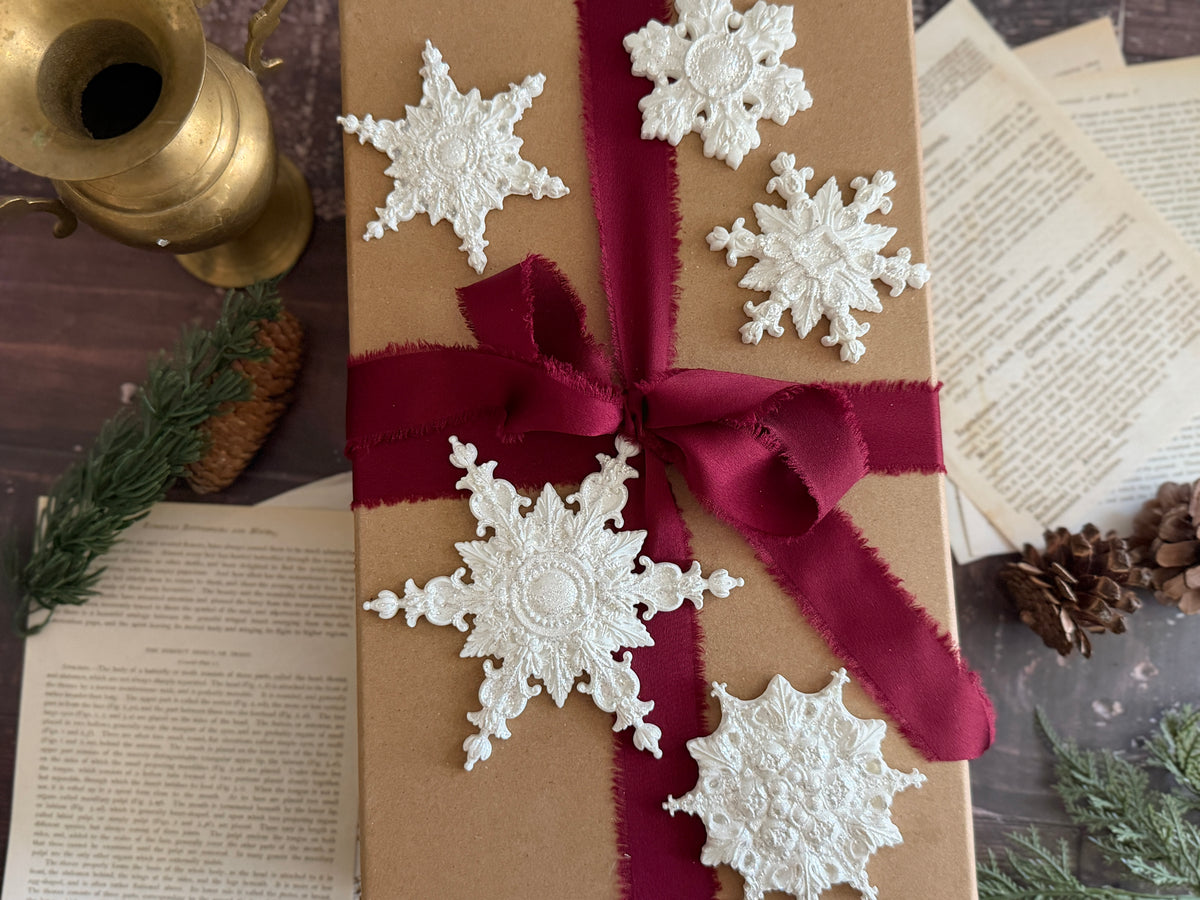 A kraft paper gift box wrapped with a deep red ribbon and bow, decorated with five white lace snowflake ornaments, styled on a dark wooden surface with vintage books, pinecones, and greenery.