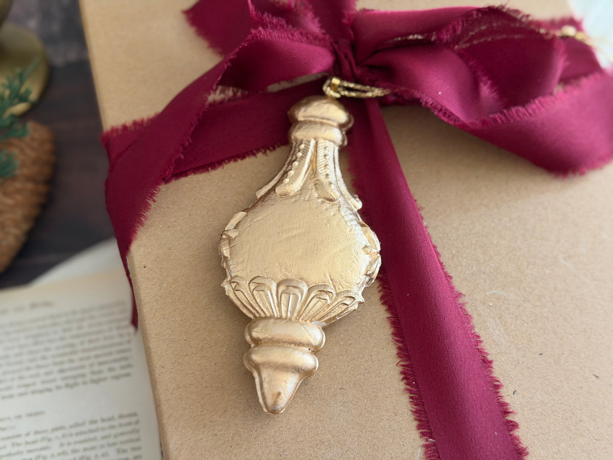 A close-up of a gold-toned decorative ornament shaped like a small bottle or vase, hanging from a deep burgundy ribbon tied around a kraft paper gift box.