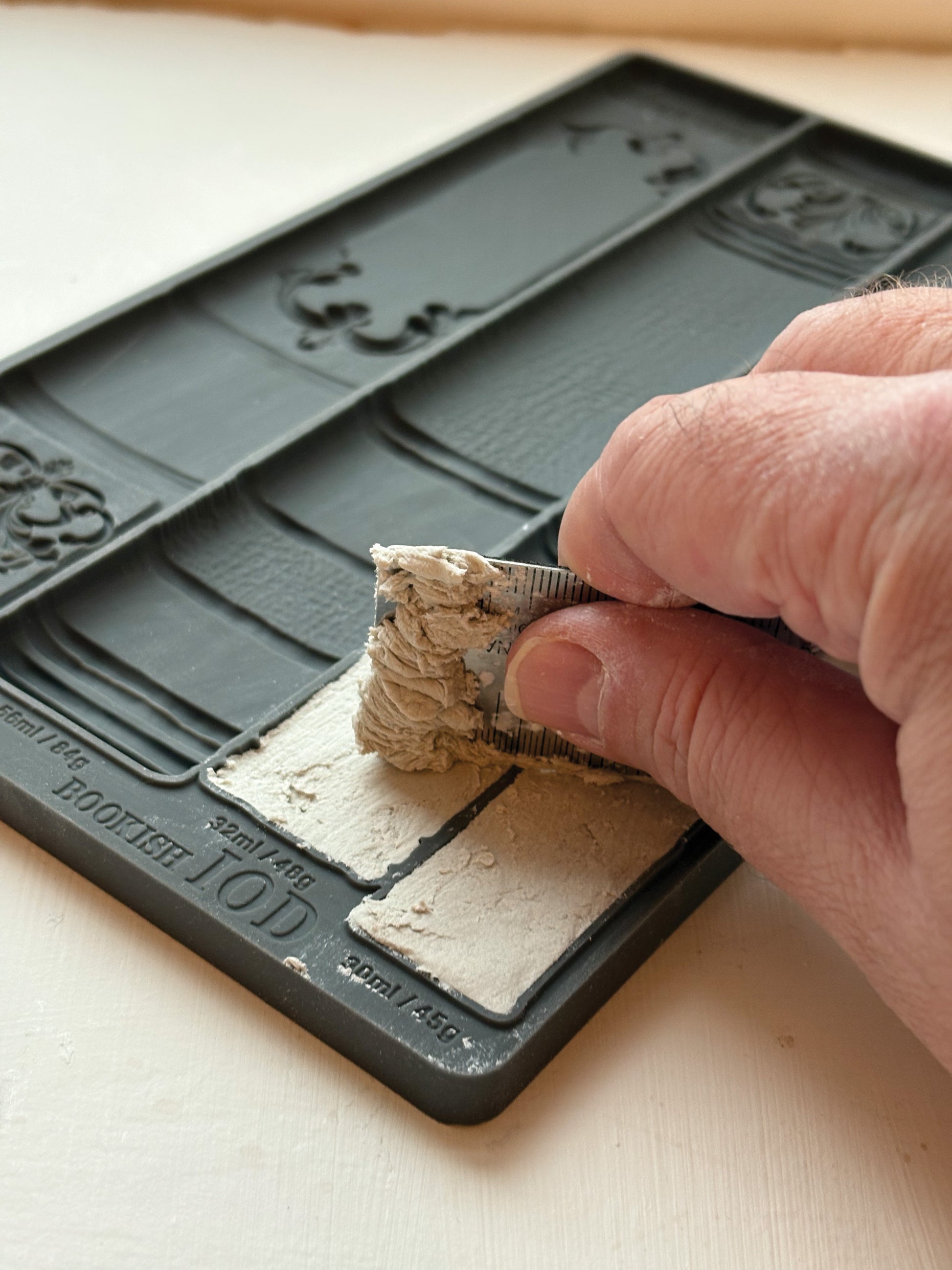 Hand using a straight edge to cast air dry clay in the Bookish mould and level it.