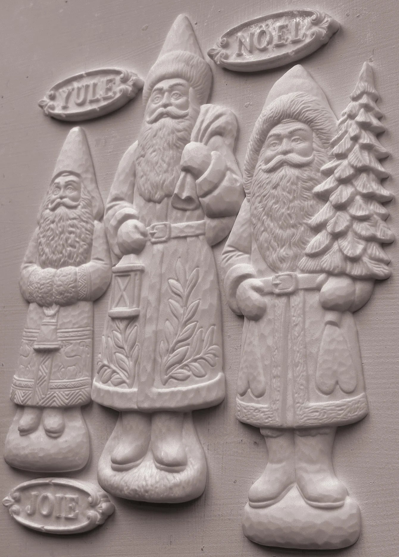 Three clay-cast Santa Claus figures in a relief style, labeled Yule, Noel, and Joy, on a textured gray background.