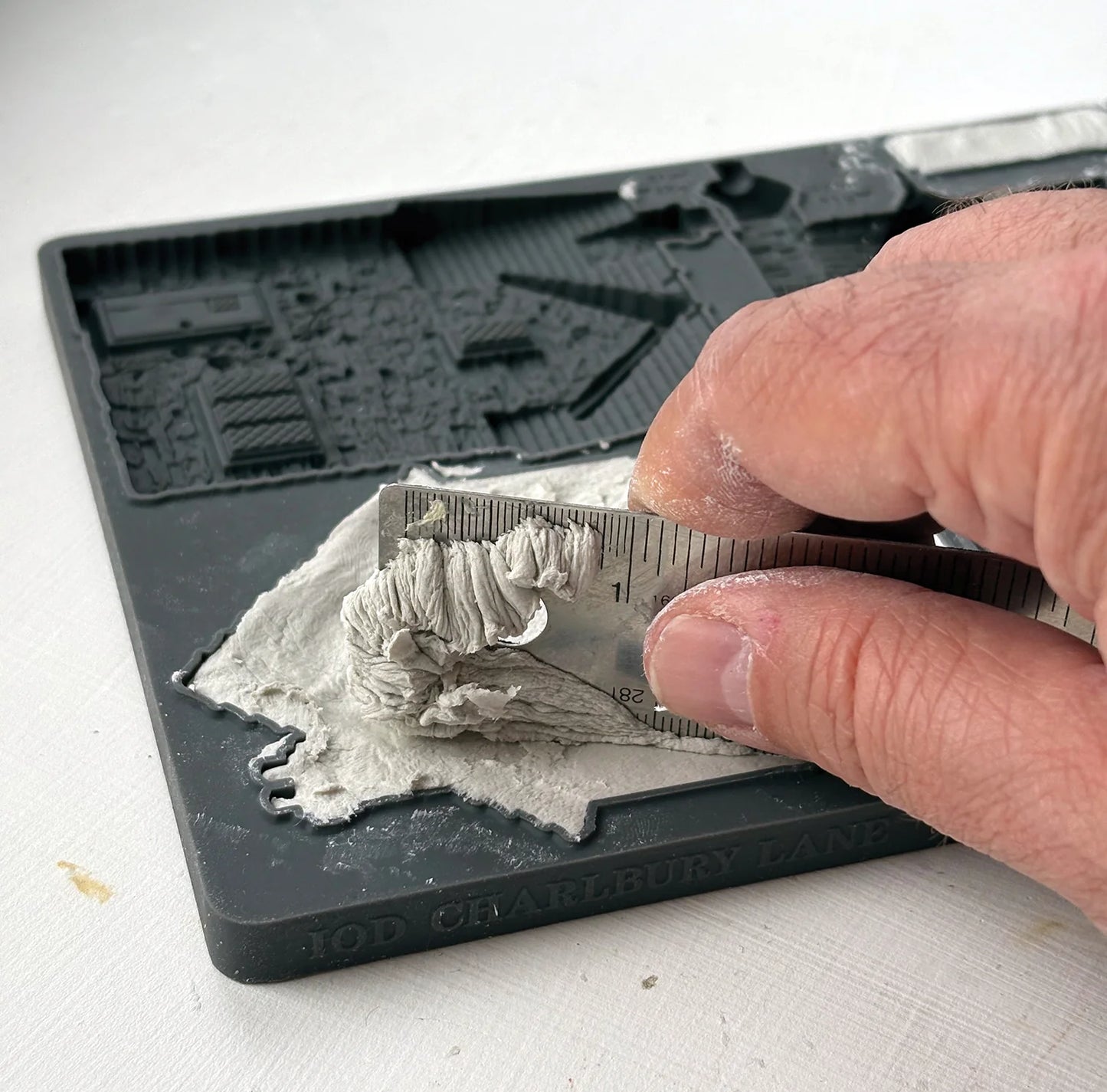 A close-up of a hand using a metal ruler to scrape excess clay from the IOD 'Charlbury' mould.
