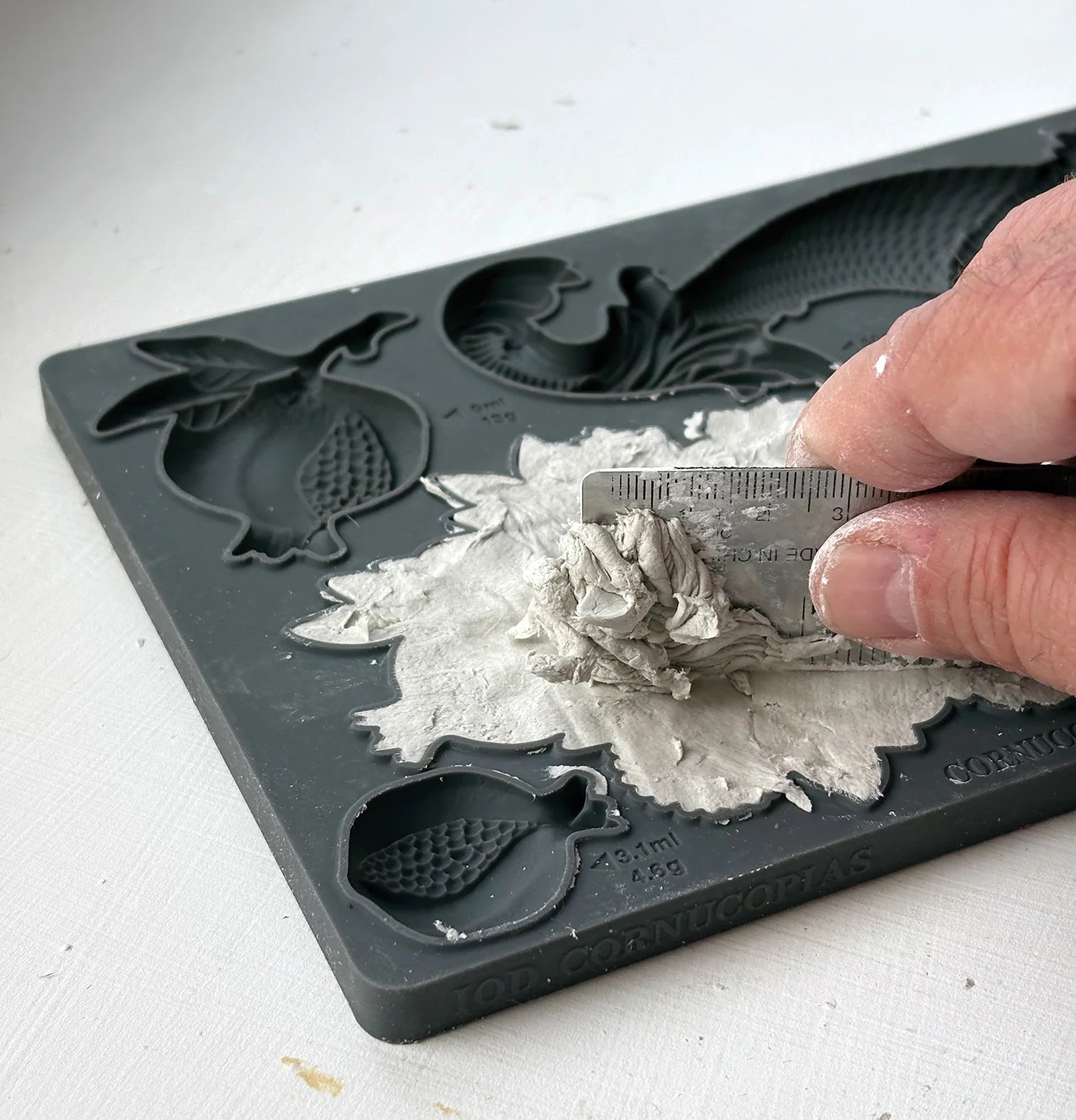 A close-up of a hand using a small metal scraper to remove clay from a dark gray silicone mold tray with fruit shaped cavities