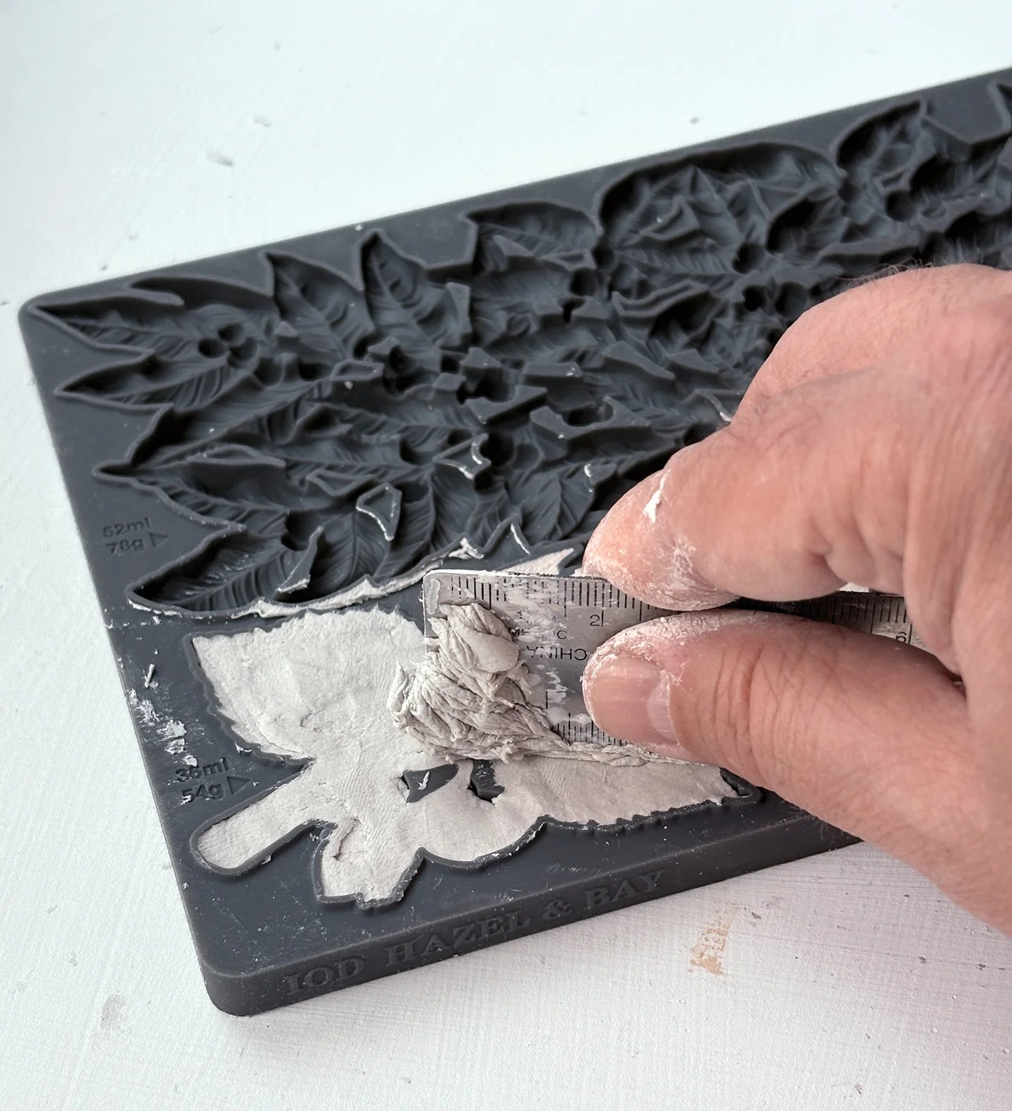 A close-up of a hand scraping excess clay out of the 'Hazel & Bay' mold, showing the mold's floral and leaf patterns.