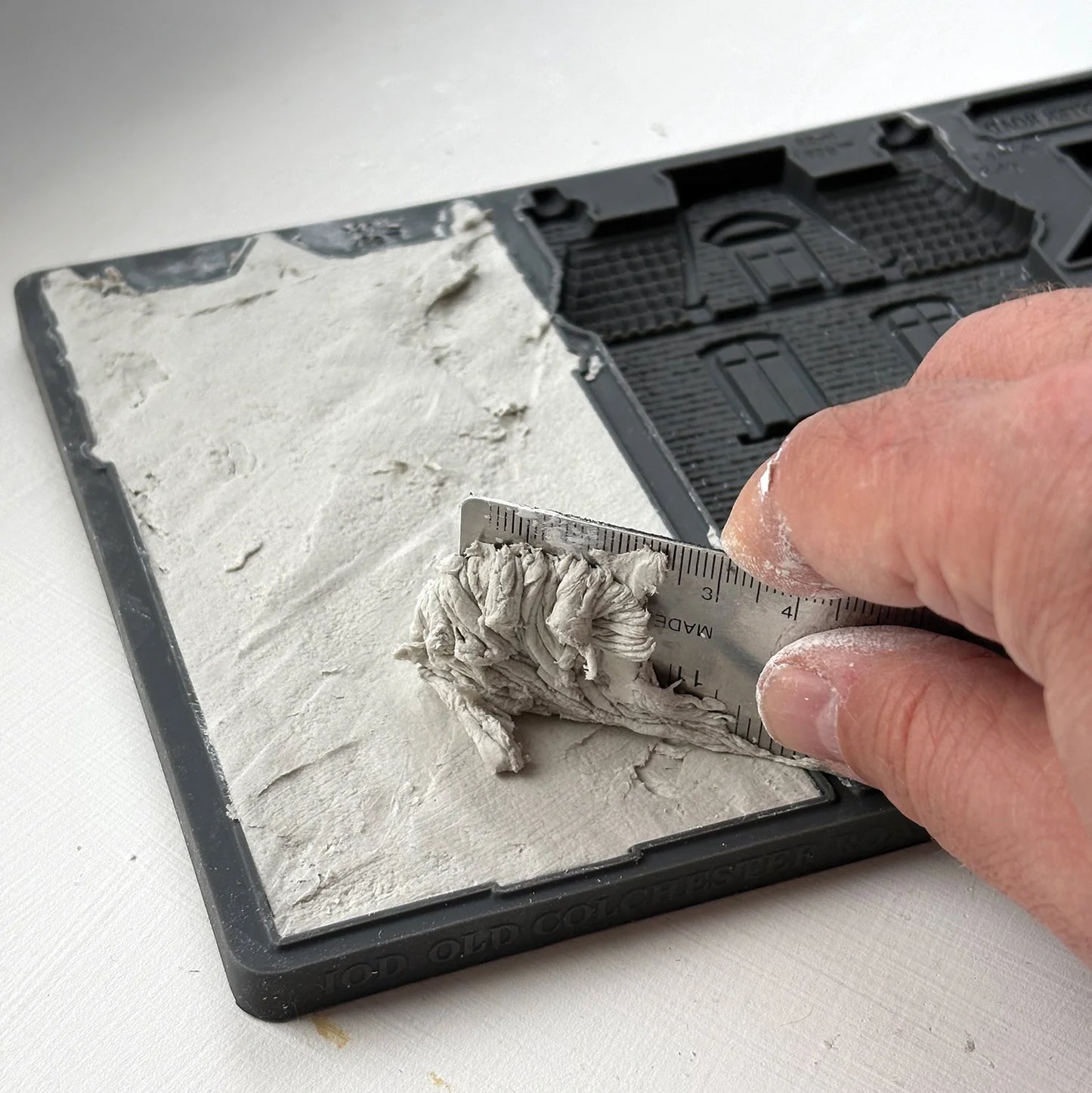 A close-up of a hand using a metal ruler to remove excess clay from a house design in the Colchester mould.