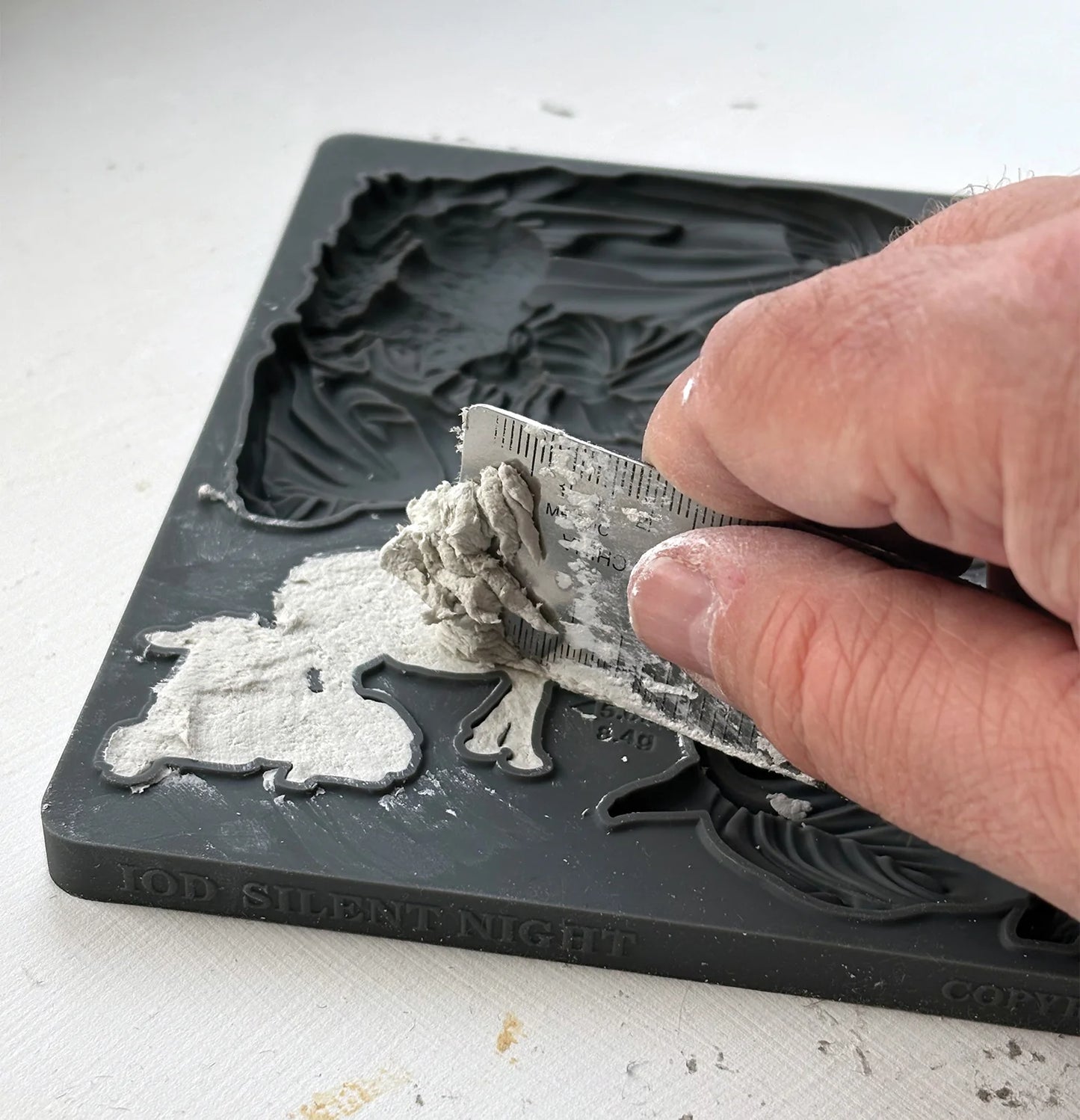 A close-up of a hand using a metal spatula to scrape excess air dry clay out of a dark gray custom-shaped silicone mold, with the embossed text 'IOD SILENT NIGHT' visible on the bottom edge.