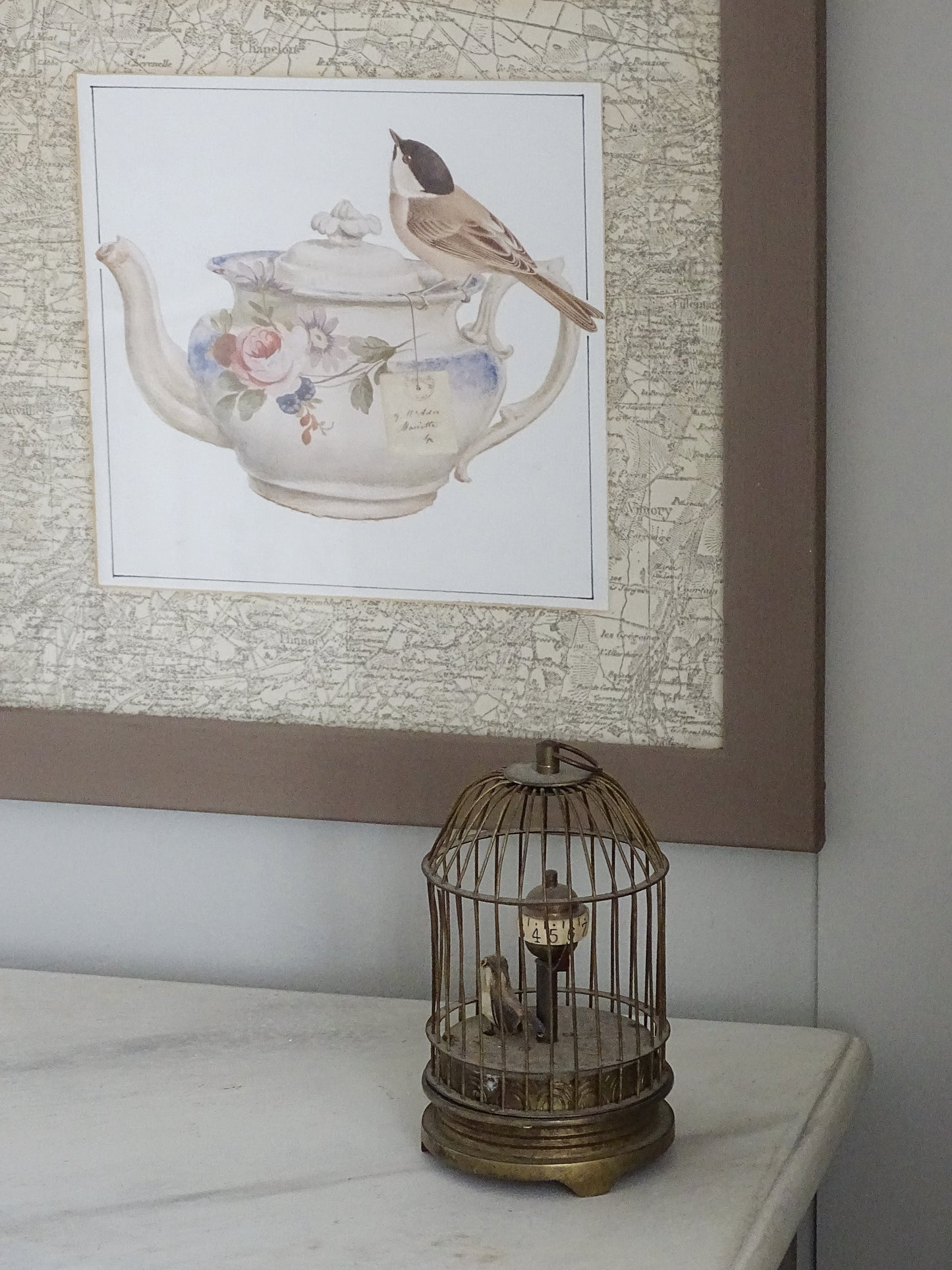 Decorative teapot with floral design and bird on a shelf, framed artwork in the background.