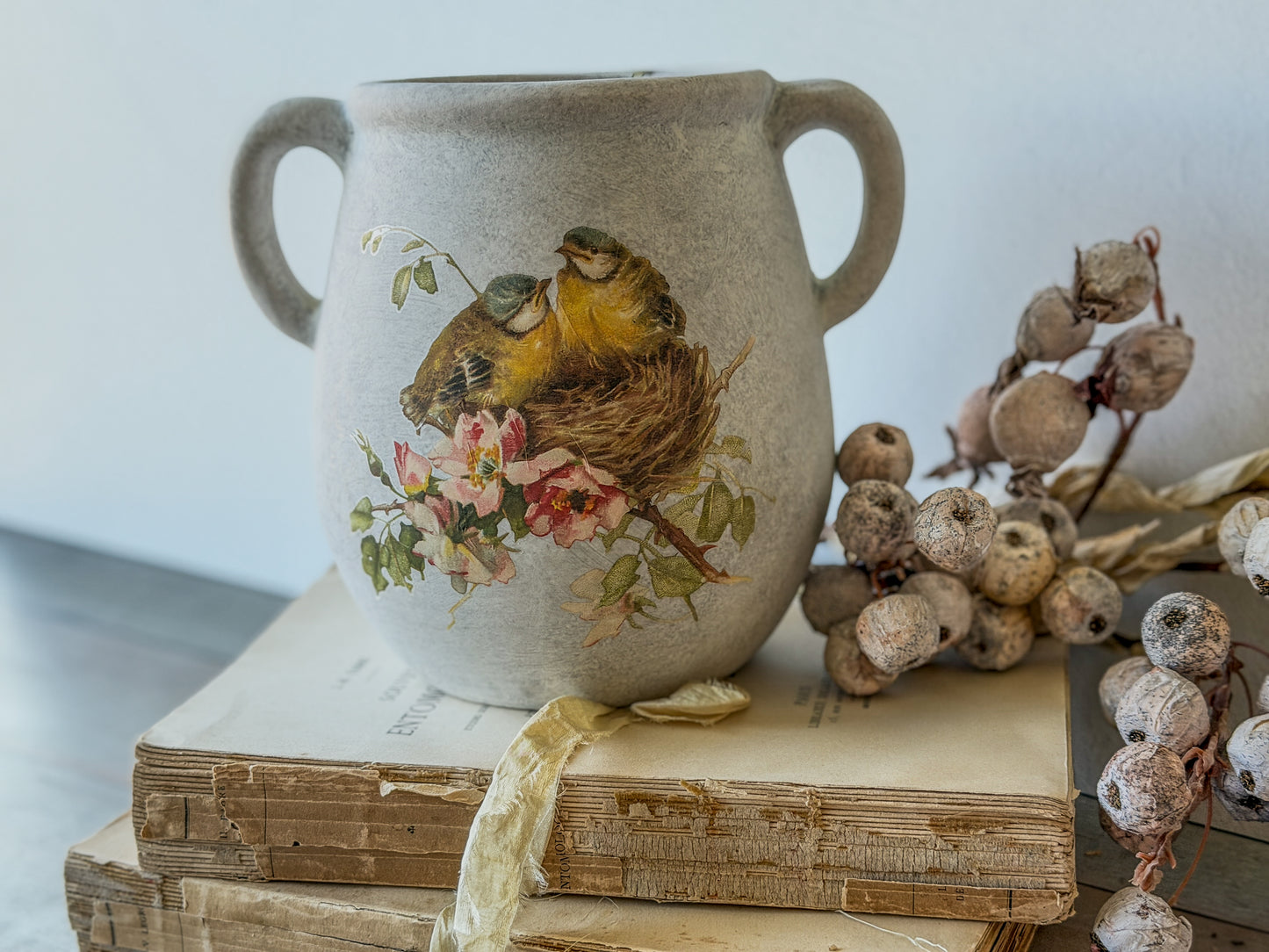 Decorative vase with bird and floral design on top of vintage books with dried berries.