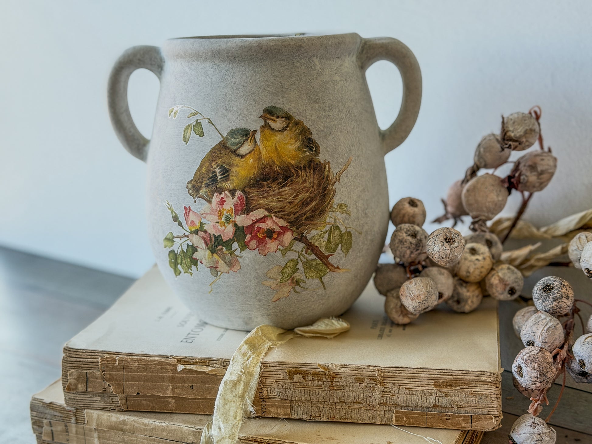 Decorative vase with bird and floral design on top of vintage books with dried berries.