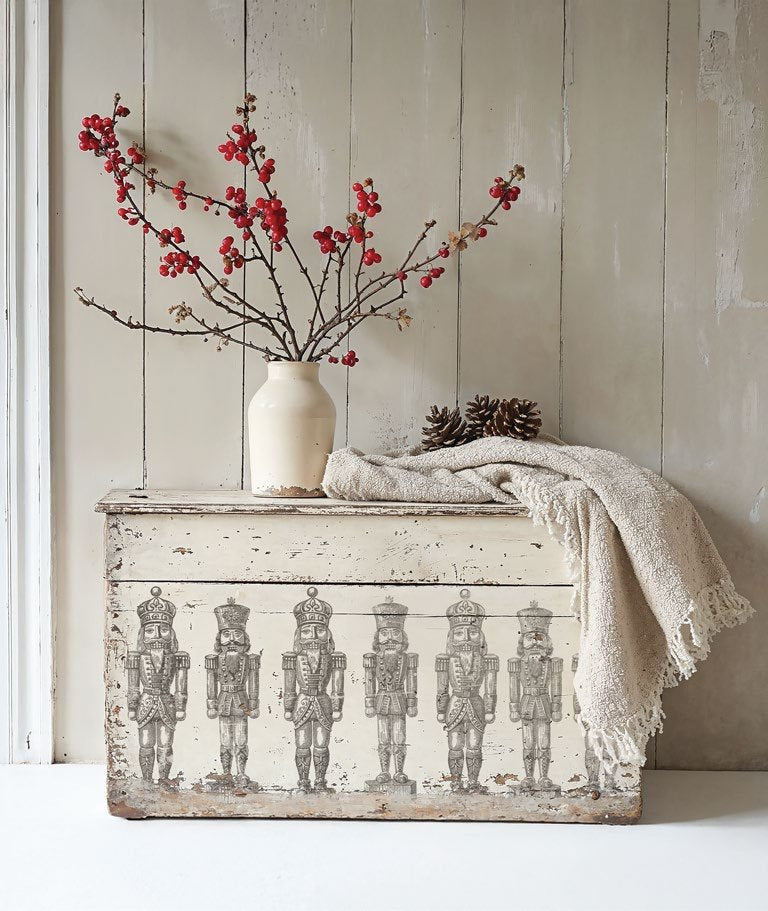 Decorative chest, stamped with the "Sugars Plum" stamp from "IOD", featuring a nutcracker design, white vase with red berries, and beige blanket against a wooden wall.