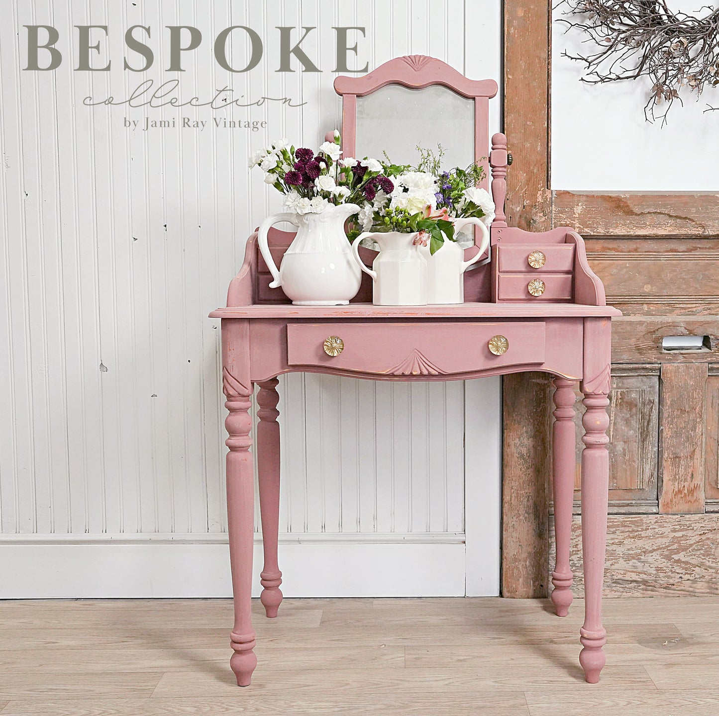 A pink vintage-style wooden dressing table in the shade "Trellis Rose" with a mirror, adorned with white vases and floral arrangements, set against a rustic backdrop.