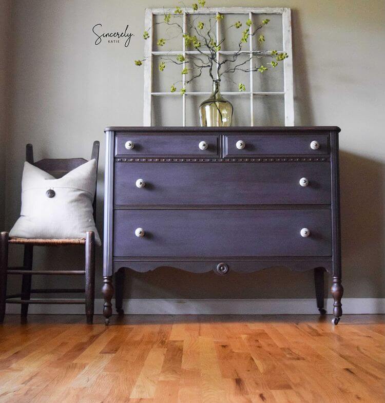 Dark purple, milk painted dresser with white knobs in a room with wooden floor and gray wall.