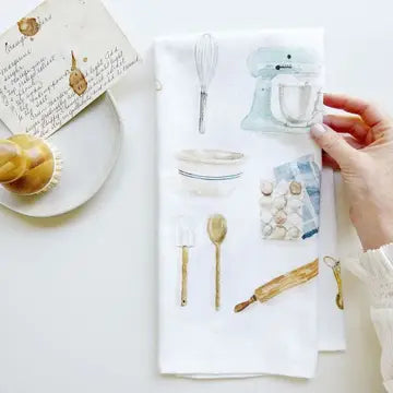 Hand holding a baking-themed tea towel with illustrations of baking tools on a white surface.