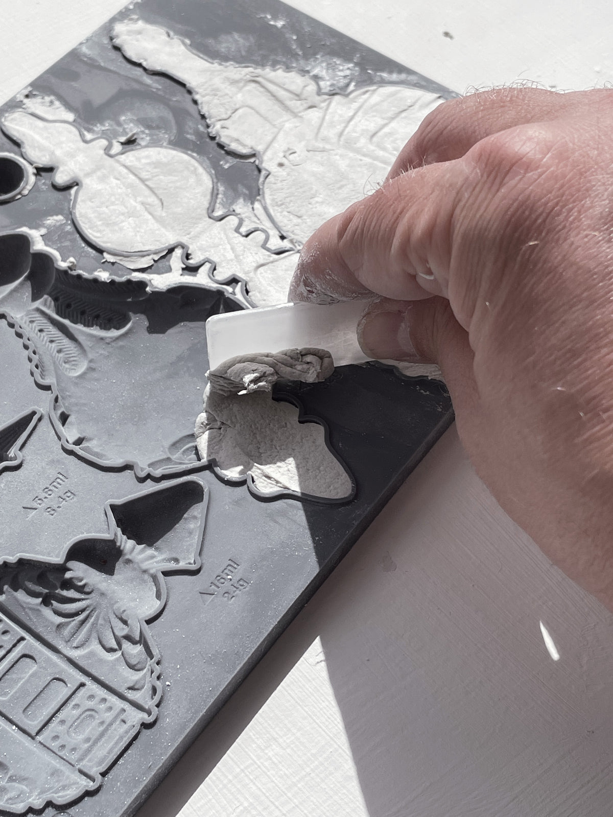 A close-up of a hand using a white spatula to scrape light gray material from a dark gray silicone mold with various decorative shapes.