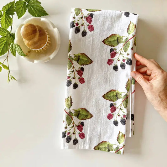 Hand holding a blackberry-patterned tea towel with berries and leaves on a white surface.