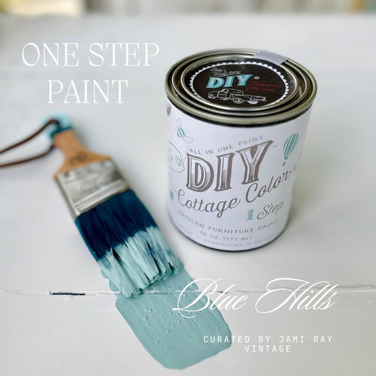 An unopened can of DIY Cottage Color paint in the shade Blue Hills, sitting on a white table with a used brush dipped in blue lying next to the can.