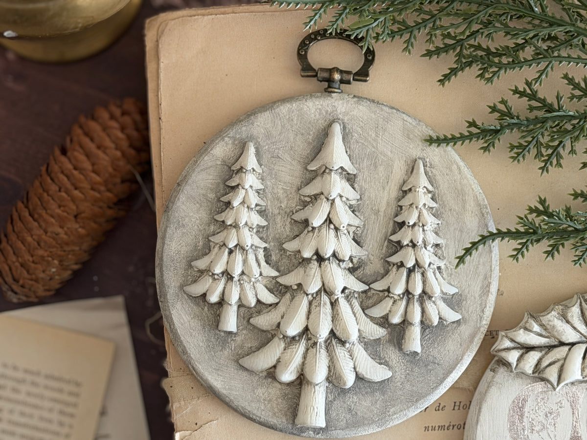 A round decorative wall plaque featuring three raised Christmas tree silhouettes, styled on a vintage book with pine branches and a pinecone nearby.