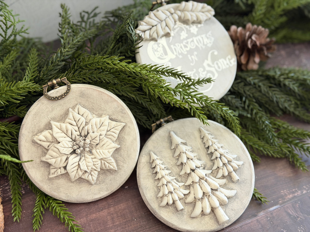 Three round beige holiday ornaments with raised relief designs, displayed on green pine branches over a dark wooden surface. One ornament features a poinsettia flower, another shows three Christmas trees, and the third has cursive text reading 'Christmas in a Song'.