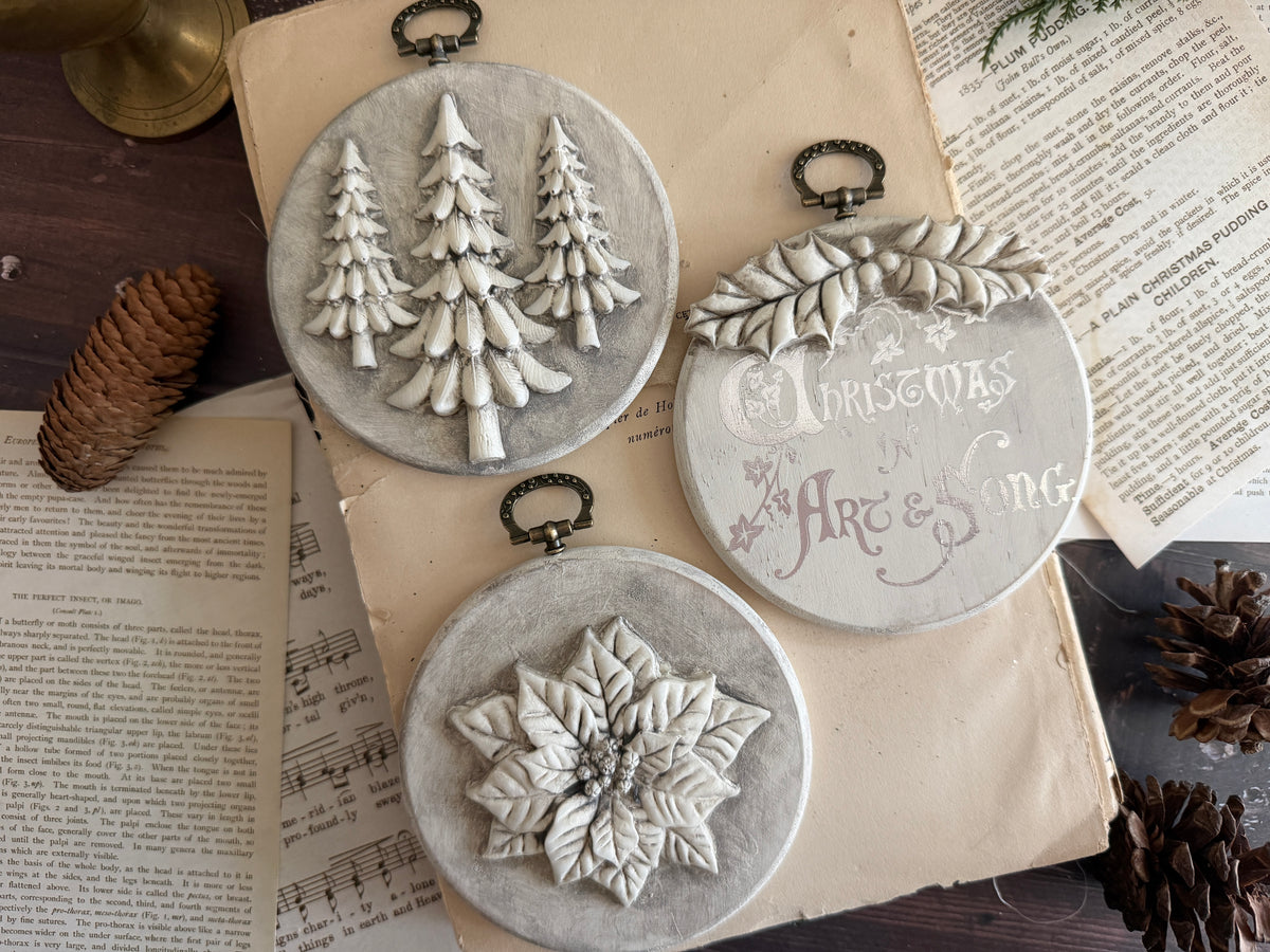 Three round gray decorative wall hangings with hanging loops, displayed on vintage songbook pages with pinecones around them. One ornament shows three white Christmas trees, another has a white floral motif, and the third reads 'Christmas is a Holiday' in white script.
