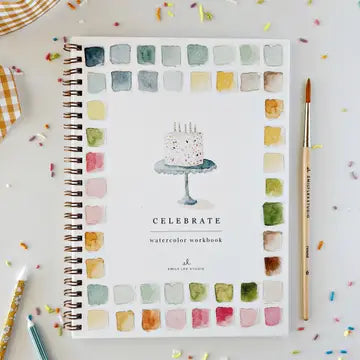 A spiral-bound watercolor workbook titled 'CELEBRATE' lies open on a white surface, surrounded by paintbrushes, confetti, and a small checkered cloth. The cover features a watercolor cake illustration and a border of multicolored paint swatches.