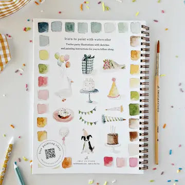 A spiral-bound watercolor painting book with party-themed illustrations and color swatches, styled on a white surface with a paintbrush, pencils, and confetti.