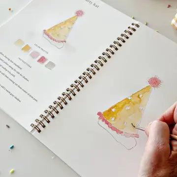 A hand is painting a watercolor party hat in a spiral-bound notebook. The notebook page shows a sample of the watercolor paper and printed text, with another finished watercolor picture of a party hat on the right.