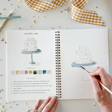 A person's hands are shown painting a watercolor cake illustration in a spiral-bound workbook. The left page shows a finished 'sprinkle cake' with text and a row of color chips, while the right page shows the hand painting.