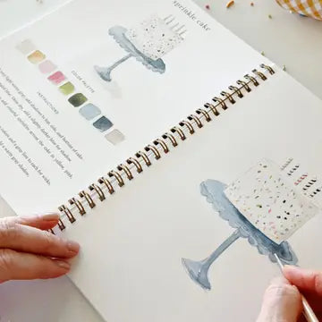 Open workbook with illustrations of a cake stand and color swatches, held by a person.