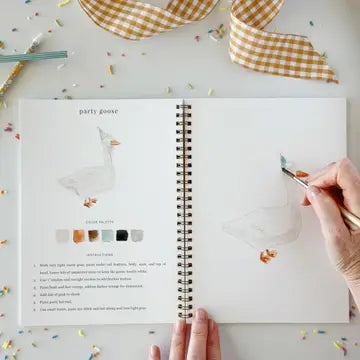 A person's hands are shown painting on a spiral-bound art journal page featuring a 'party goose' design. The scene is styled as a flat-lay with a yellow and white gingham ribbon, paintbrushes, and confetti on a white surface.