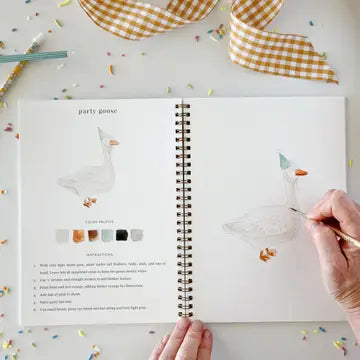 An open spiral-bound watercolor workbook on a white surface, showing illustrated goose designs, color palettes, and instructions. A hand is coloring one goose with a gold pen, and another hand holds the bottom edge of the page.