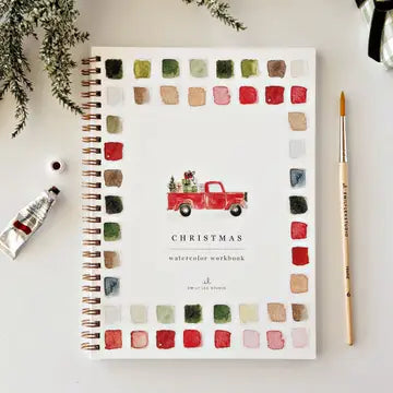 A spiral-bound watercolor workbook titled 'CHRISTMAS watercolor workbook' by Emily Lex Studio, styled in a flat lay with a red truck illustration and watercolor swatches around the border. A paint tube, paintbrush, and greenery are arranged around it on a light background.