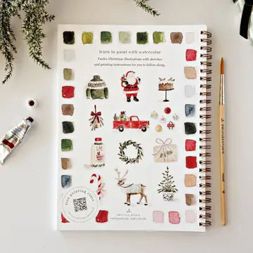 A spiral-bound watercolor painting book titled 'Learn to paint with watercolor' featuring Christmas illustrations, styled on a white surface with a paint tube, brush, and greenery.