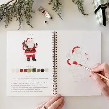 An open spiral-bound watercolor journal laid on a white surface, showing a Santa illustration with color palette and instructions on the left page, and a partially painted outline of Santa on the right page. A hand holds a paintbrush over the right page, and holiday greenery, a tube of paint, and a checkered fabric corner are visible around it.