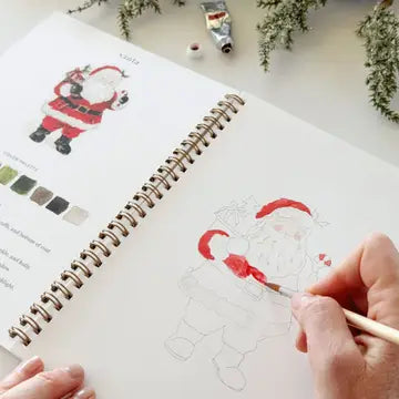 A hand uses a fine paintbrush to watercolor paint a Santa illustration in a spiral-bound workbook. The page shows a color palette and another completed Santa drawing, with a tube of paint and greenery on a light tabletop.