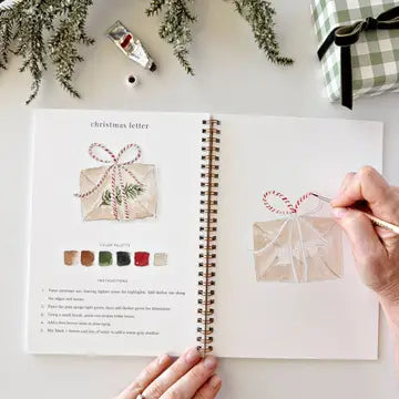 An open spiral-bound art journal shows a Christmas letter craft design with illustrated wrapped envelopes, color swatches, and instructions. Hands are visible holding the pages, and holiday greenery, a paint tube, and a wrapped gift appear around it on a white surface.