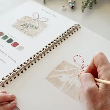 A hand holding a watercolor paintbrush paints a Christmas letter envelope with red-and-white striped ribbon on a spiral-bound art journal. The open page shows a finished example illustration and a small color palette.