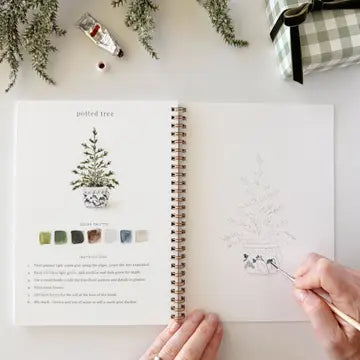 An open spiral-bound watercolor workbook on a white surface, showing a potted tree illustration and instructions on one page, with a hand holding a paintbrush working on a matching sketch on the other page. A small tube of paint, greenery, and a wrapped gift are visible around it.