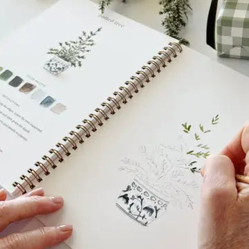 A watercolor  journal page featuring a potted tree illustration, held open by two hands. The page has a spiral binding and is shown on a light surface with a small green-and-white checkered box in the background.