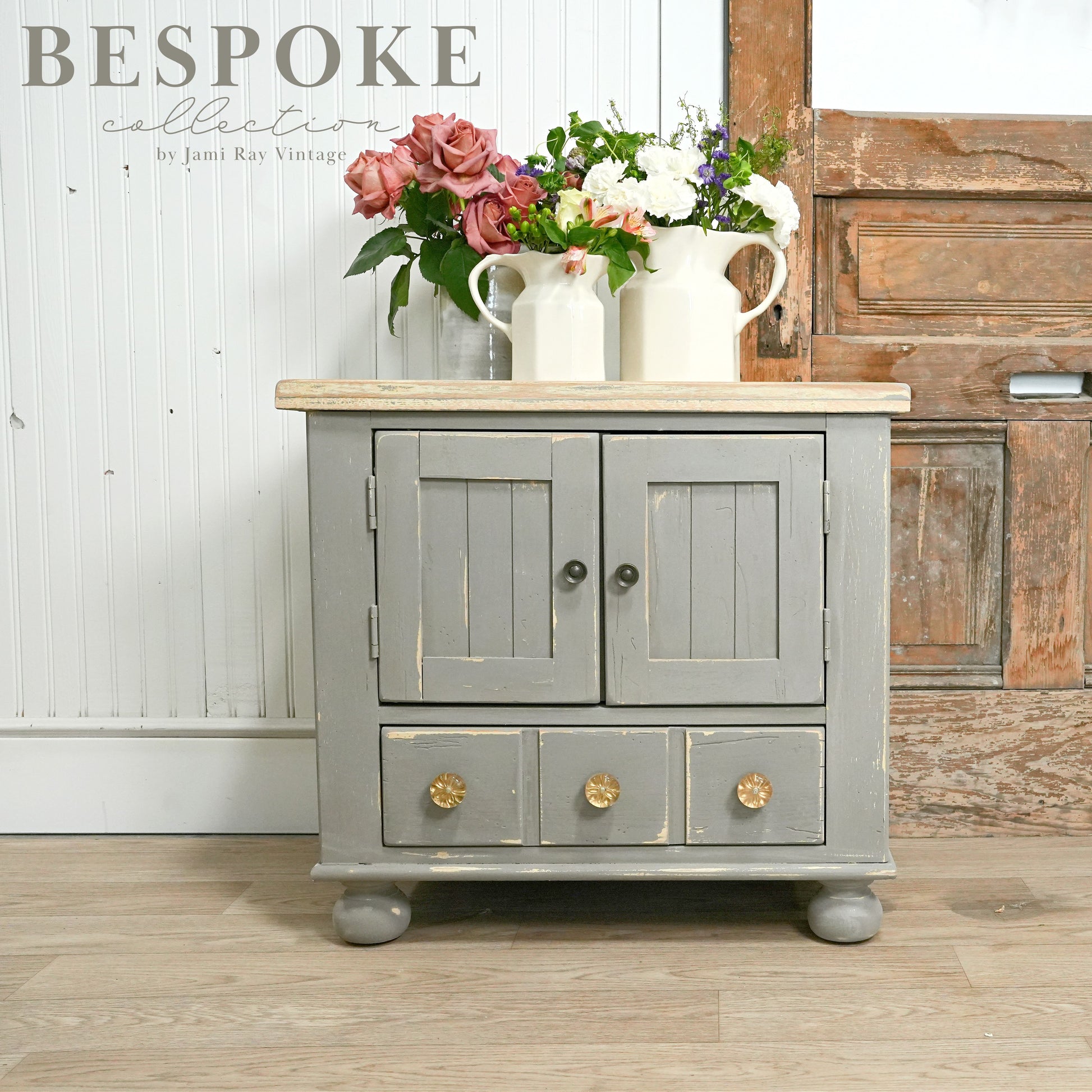 A small wooden cabinet painted in a neutral cobblestone color with a distressed finish, featuring gold handles and a natural wood top, placed against a rustic backdrop with a vase of flowers on top.