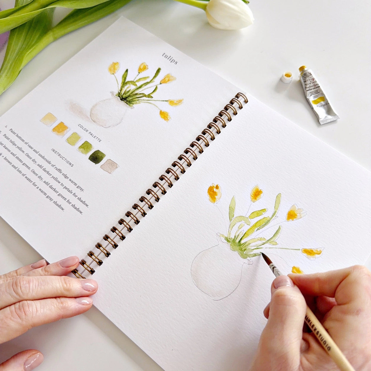 Person watercolor painting flowers in a notebook with a paintbrush, surrounded by art supplies on a white surface.