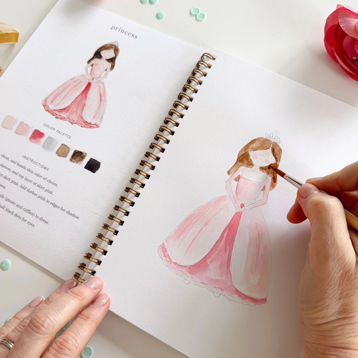Person watercolor painting a princess in a pink dress in a watercolor workbook with color swatches.