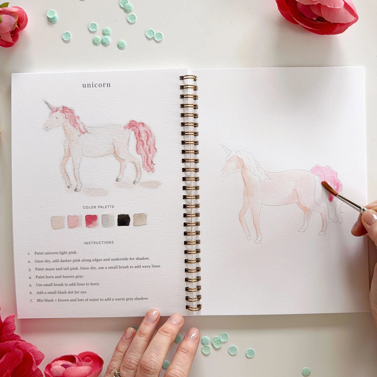 Open book with unicorn illustration and color palette, surrounded by flowers.